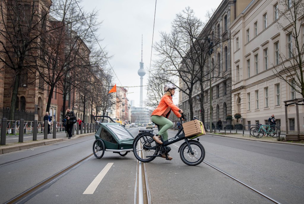 Eine Frau fährt auf einem E-Lastenrad mit Kinderanhänger quer über eine Straße. Im Hintergrund ist der Berliner Fernsehturm zu erkennen.