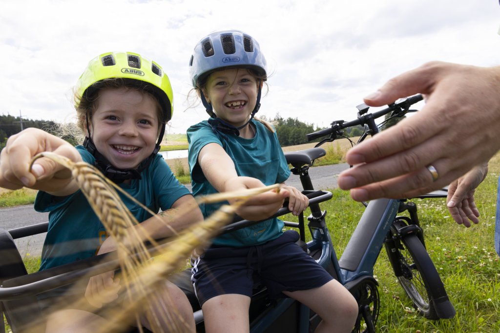 Zwei Kinder sitzen auf Fahrrad, haben einen Fahrradhelm auf und Getreide in der Hand.