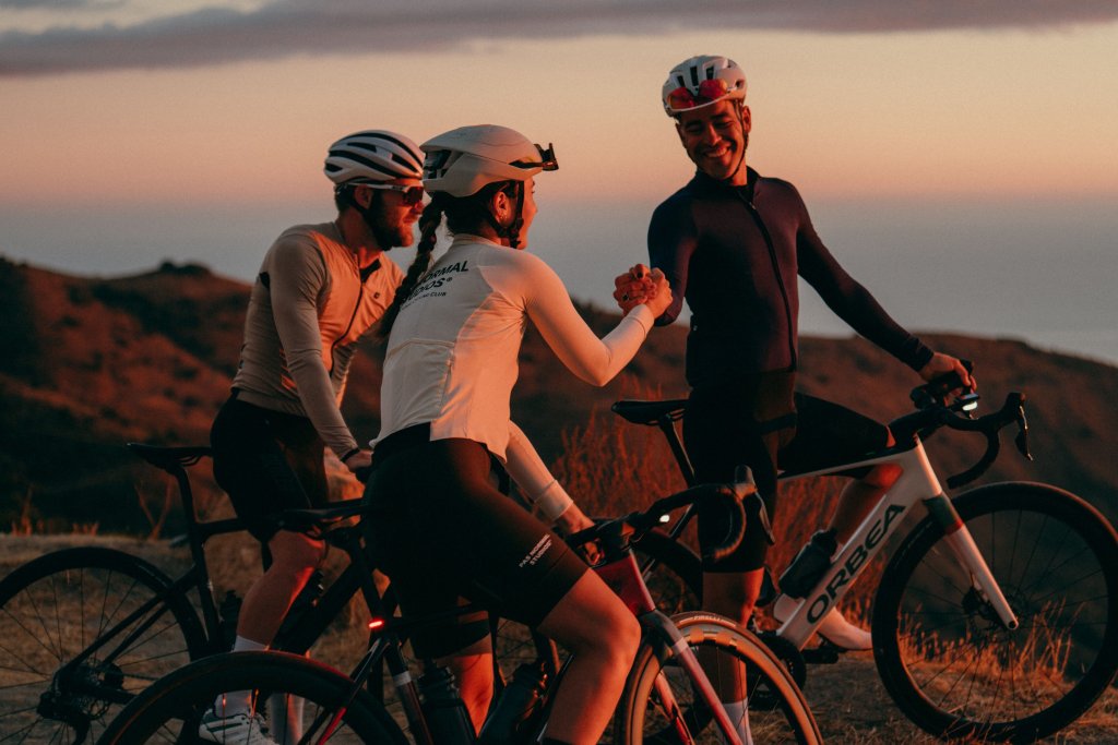 Drei Personen stehen mit Rennrädern im Abendlicht auf einem Hügel und zwei geben sich die Hand.